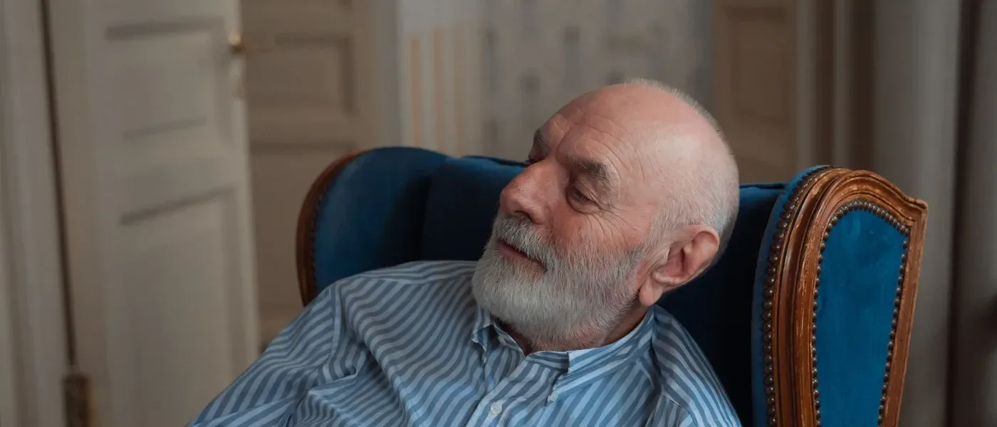 Older man on an armchair holding a TV remote — a common warning sign of hearing loss is quietly turning up the volume year after year